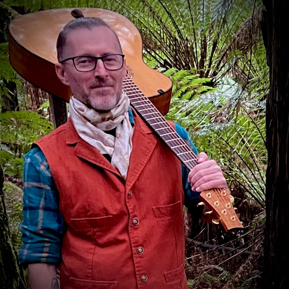 Edward Bassingthwaighte in a rust vest and scarf, an acoustic guitar resting on his shoulder, standing among tree ferns in the forest
