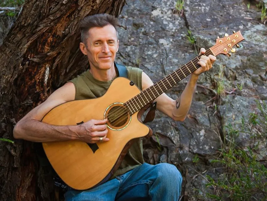 Smiling man in a green singlet holding an acoustic guitar, leaning against a tree trunk and rock face